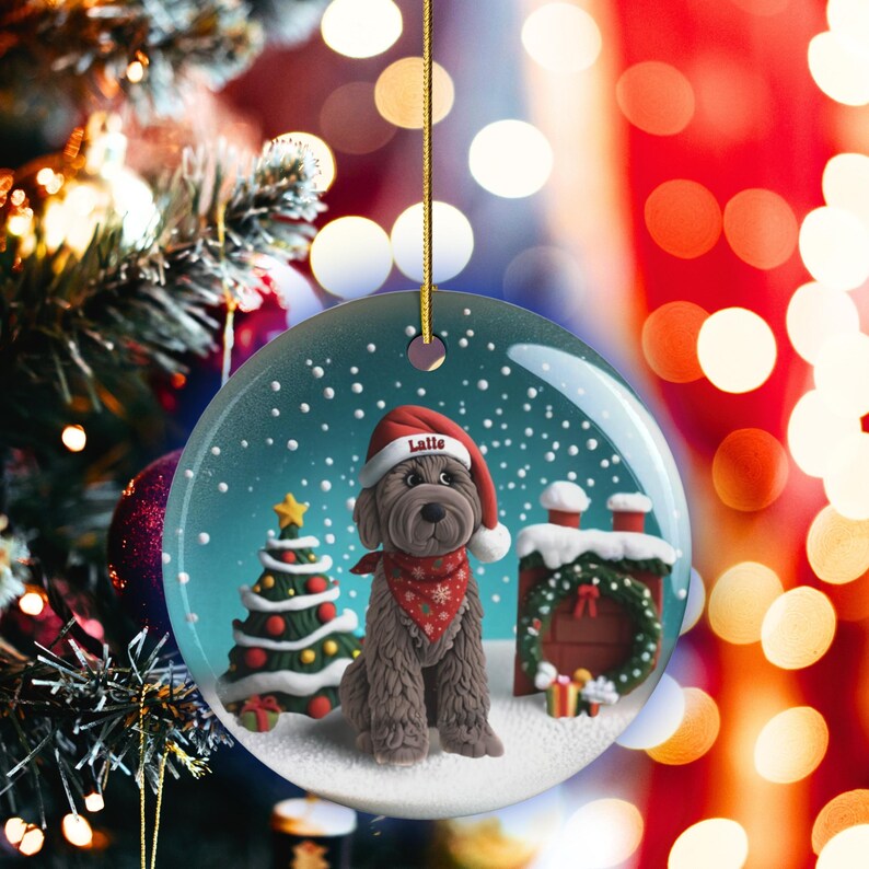 May include: A round Christmas ornament featuring a brown dog wearing a Santa hat with the name "Latte" on it, a red bandana, and a snowy scene. A Christmas tree and a chimney are also visible. The ornament hangs from a gold string.