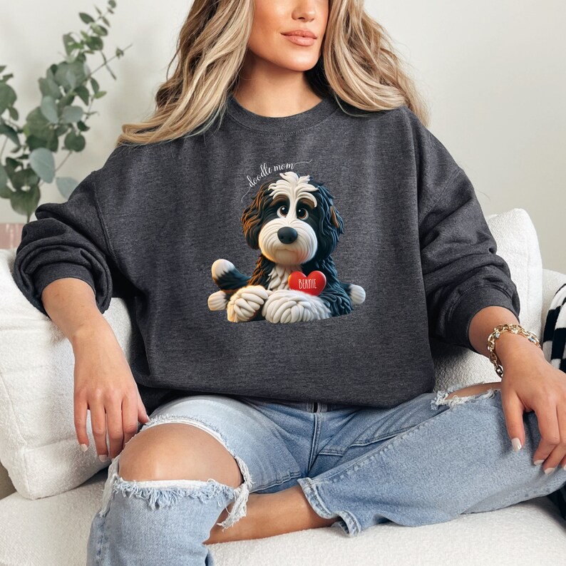 A woman wearing a Bernedoodle sweatshirt with cute design on the front and the back.  The front features a bernedoodle dog in claymation design holding a heart with his name, across the top of his head the words, "Doodle Mom".