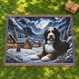 Puede incluir: Una manta tejida que presenta a un perro de monta&ntilde;a de Berna descansando frente a un pueblo invernal nevado. El fondo muestra caba&ntilde;as de madera iluminadas, pinos cubiertos de nieve y monta&ntilde;as bajo una luna llena.