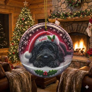 May include: A round Christmas ornament featuring a black dog wearing a Santa hat with the name "Teddi" on it. The ornament has a snowy background with holly and berries. The background shows a Christmas tree and a fireplace.