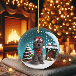 May include: A round Christmas ornament featuring a brown dog wearing a Santa hat and a red bandana. The ornament has a snowy scene with a Christmas tree and a small house. The word "Latte" is on the hat.
