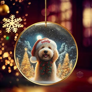May include: A round Christmas ornament with a gold rim, featuring a fluffy dog wearing a Santa hat with the name "Goldie". The dog is also wearing a festive green and red garland. The background shows a snowy winter scene with Christmas trees.
