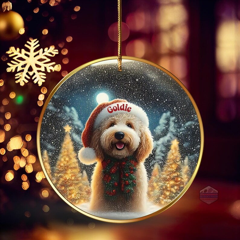 May include: A round Christmas ornament with a gold rim, featuring a fluffy dog wearing a Santa hat with the name "Goldie". The dog is also wearing a festive green and red garland. The background shows a snowy winter scene with Christmas trees.