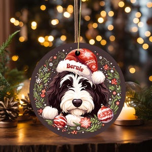 Puede incluir: Adorno navideño redondo con un perro blanco y negro con un gorro de Papá Noel rojo con el nombre "Bernie". El adorno está decorado con una corona de hojas verdes, bayas rojas y copos de nieve, y dos bolas rojas y blancas.