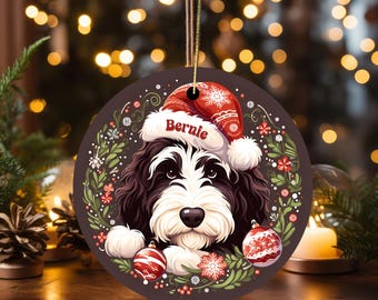 May include: Round Christmas ornament featuring a black and white dog wearing a red Santa hat with the name "Bernie". The ornament is decorated with a wreath of green leaves, red berries, and snowflakes, and two red and white striped ornaments.