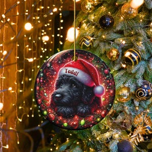 May include: Round Christmas ornament featuring a black dog wearing a Santa hat with the name "Teddi" on it. The ornament has a festive design with red, green, and white colors. It is hanging from a gold string, with a Christmas tree in the background.