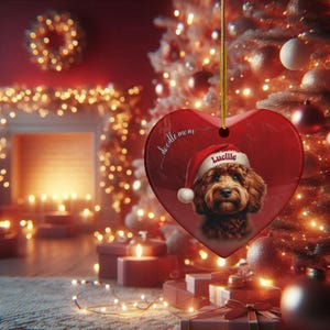 May include: A heart-shaped Christmas ornament with a red background, featuring a brown dog wearing a Santa hat. The ornament has the name "Lucille" and the words "doodle mom" on it. The ornament is hanging in front of a Christmas tree.