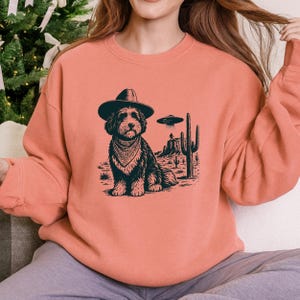 May include: Coral-colored sweatshirt featuring a black and white graphic of a dog wearing a cowboy hat and bandana, set against a desert landscape with a UFO. The dog is the central focus, with cacti and a flying saucer adding detail.