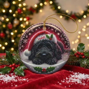 May include: A round Christmas ornament featuring a black dog wearing a Santa hat with the name "Teddi". The ornament has a gold string for hanging and is decorated with holly and snowflakes. The background is a blurred image of Christmas lights.