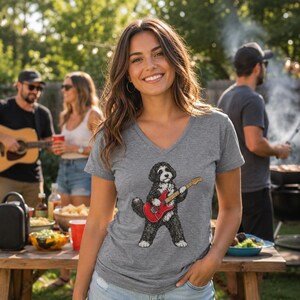 May include: A heather gray V-neck t-shirt featuring a graphic illustration of a black and white dog playing a red electric guitar. The dog has a shaggy coat and is standing on its hind legs while holding the guitar with its front paws.