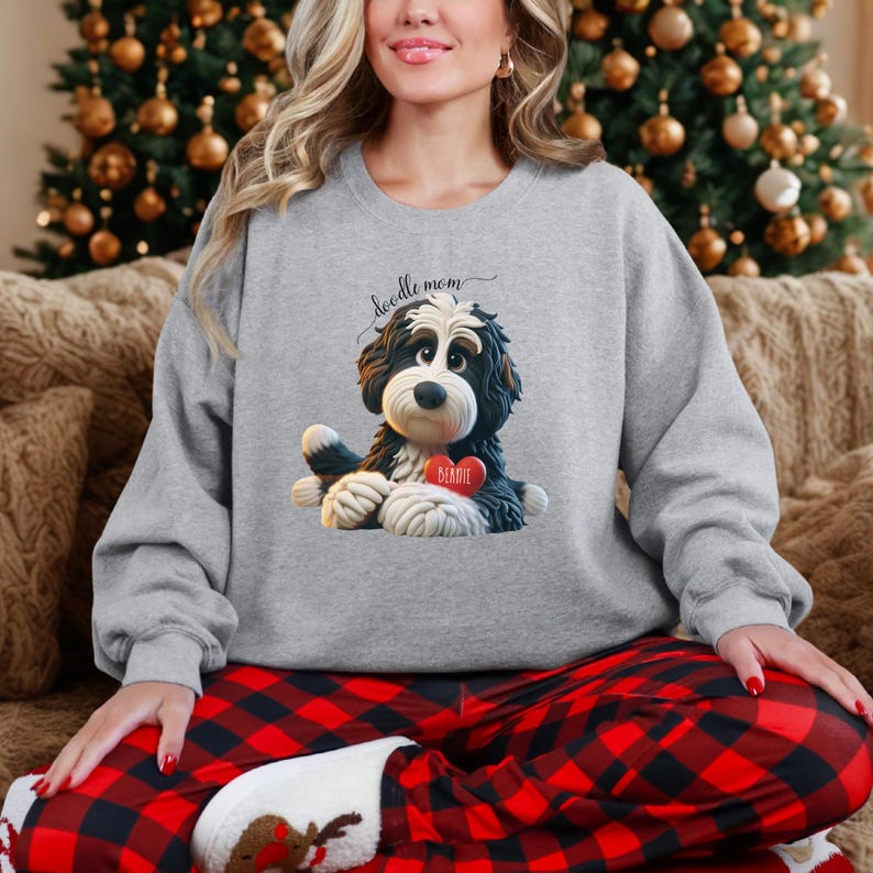 A woman wearing a Bernedoodle sweatshirt with cute design on the front and the back.  The front features a bernedoodle dog in claymation design holding a heart with his name, across the top of his head the words, "Doodle Mom".