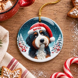 May include: A round ceramic Christmas ornament featuring a Bernese Mountain Dog wearing a red Santa hat with the name Bernie printed on the brim. The dog is set against a snowy winter landscape with pine trees and snowmen.