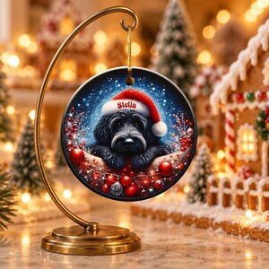 May include: A round ceramic Christmas ornament hanging from a gold metal stand. The ornament features a black dog wearing a red Santa hat with the name Stella printed on the brim, surrounded by red berries and ornaments on a blue background.