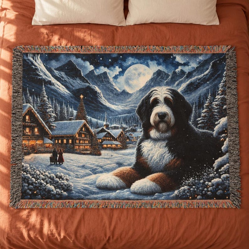 Puede incluir: Una manta tejida rectangular que presenta a un gran perro de monta&ntilde;a de Berna tumbado en primer plano. El fondo muestra un pueblo nevado de noche con casas iluminadas, pinos, monta&ntilde;as y una luna llena en el cielo.