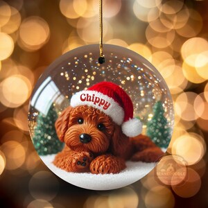 May include: A clear, round Christmas bauble featuring a brown dog wearing a red Santa hat with the name "Chippy" on it. The bauble has a snowy base with small green trees and a gold string for hanging. The background is a bokeh of warm, golden lights.