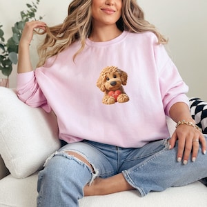 May include: A light pink sweatshirt featuring a cartoon illustration of a fluffy, brown dog holding a red heart. The dog has a sweet expression. The sweatshirt is a solid color and has long sleeves.
