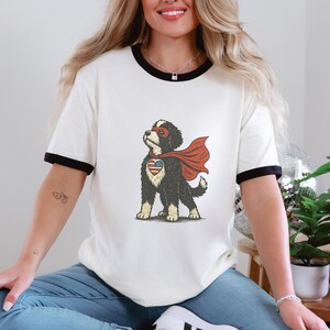 May include: Cream-coloured t-shirt with black trim featuring a cartoon superhero dog with a red cape, mask, and a heart-shaped emblem of the American flag. The dog is black and white.
