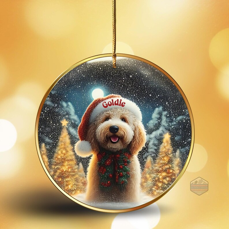 May include: A round Christmas ornament with a gold rim, featuring a dog wearing a Santa hat with the name "Goldie" on it. The dog has a red and green garland around its neck, set against a snowy winter scene with Christmas trees.
