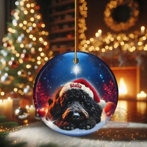 May include: Round Christmas ornament featuring a black dog wearing a Santa hat with the name "Stella" on it. The ornament has a blue background with a star and snow. The ornament is hanging from a gold string.