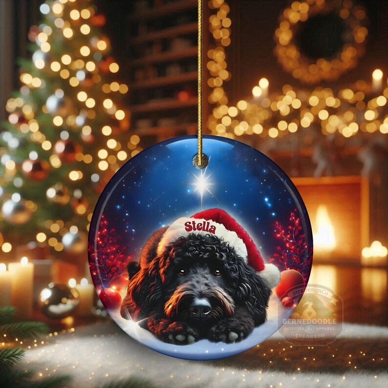 May include: Round Christmas ornament featuring a black dog wearing a Santa hat with the name "Stella" on it. The ornament has a blue background with a star and snow. The ornament is hanging from a gold string.