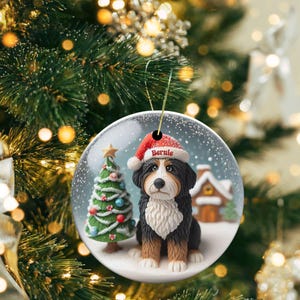 May include: A round Christmas ornament featuring a Bernese Mountain Dog wearing a Santa hat with the name "Bernie". The ornament has a snowy scene with a Christmas tree and a small house. The ornament is hanging from a gold string.