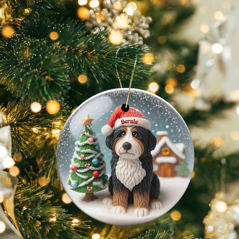 May include: A round Christmas ornament featuring a Bernese Mountain Dog wearing a Santa hat with the name "Bernie". The ornament has a snowy scene with a Christmas tree and a small house. The ornament is hanging from a gold string.
