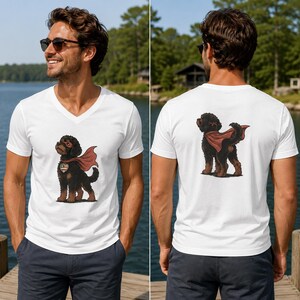 May include: A white V-neck t-shirt featuring a graphic of a black curly-haired dog wearing a red superhero cape and a heart-shaped tag on the front, and the same dog viewed from the back on the rear of the shirt, showcasing the cape detail.