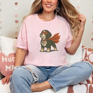 May include: A light pink t-shirt featuring a cartoon dog dressed as a superhero. The dog has a red mask, cape, and a heart-shaped emblem with the American flag. The shirt is a casual style.
