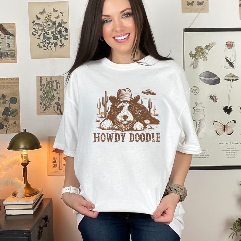 Może przedstawiać: Biała koszulka z brązową grafiką psa w kapeluszu kowbojskim i bandanie, z kaktusami i UFO. Tekst "HOWDY DOODLE" znajduje się poniżej. Koszulka ma luźny kr&oacute;j.