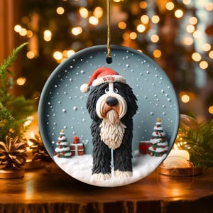 May include: A round Christmas ornament featuring a black and white dog wearing a red Santa hat with the name "Willy". The ornament has a snowy scene with Christmas trees and presents. The ornament is hanging by a gold string.