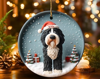May include: A round Christmas ornament featuring a black and white dog wearing a red Santa hat with the name "Willy". The ornament has a snowy scene with Christmas trees and presents. The ornament is hanging by a gold string.