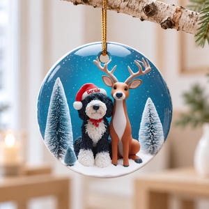 May include: A round ceramic Christmas ornament featuring a black and white dog wearing a red Santa hat with the name Bernie printed on it, sitting next to a brown deer in a snowy forest with blue background and white pine trees.
