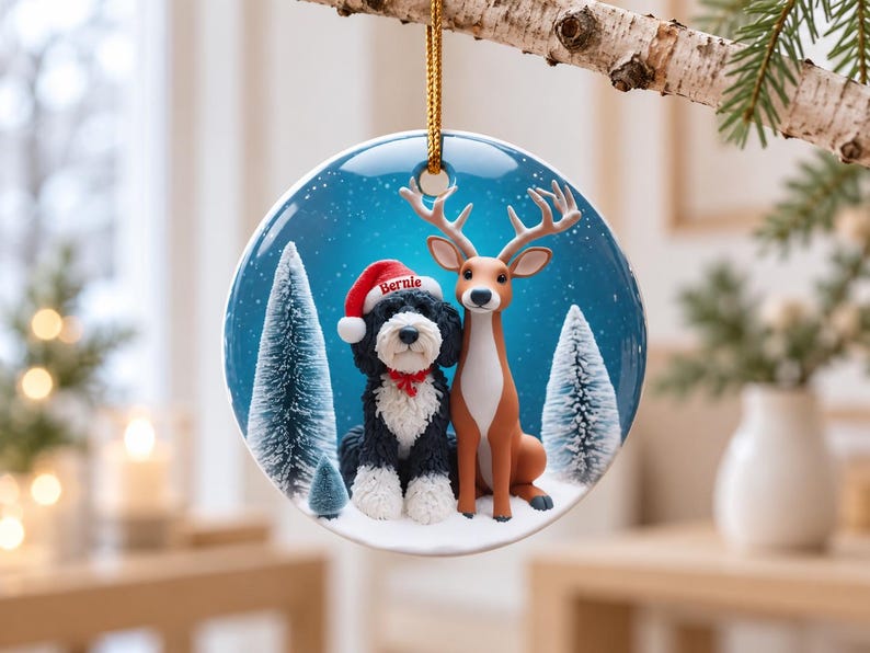 May include: A round ceramic Christmas ornament featuring a black and white dog wearing a red Santa hat with the name Bernie printed on it, sitting next to a brown deer in a snowy forest with blue background and white pine trees.