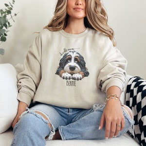 Puede incluir: Sudadera beige con un gráfico de un perro Bernedoodle con el texto "doodle mom" arriba y "BERNIE" abajo. El perro tiene pelo negro, marrón y blanco. La sudadera es de corte holgado.