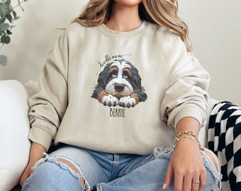 May include: Beige sweatshirt featuring a graphic of a Bernedoodle dog with the text "doodle mom" above and "BERNIE" below. The dog has black, brown, and white fur. The sweatshirt is a relaxed fit.