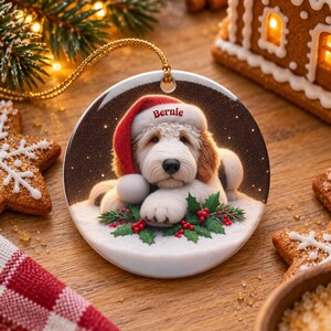 May include: A round ceramic Christmas ornament featuring a fluffy white and tan dog wearing a red Santa hat with the name Bernie printed on the brim. The dog rests on snow with green holly leaves and red berries, suspended by a gold cord.