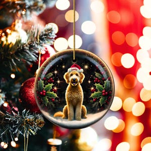 May include: A round Christmas ornament featuring a golden doodle dog wearing a Santa hat with the name "Goldie" on it. The ornament has a black background with holly and berries, and is hanging from a gold string.