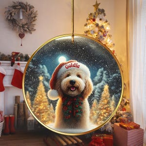 May include: A round Christmas ornament featuring a fluffy dog wearing a Santa hat with the name "Goldie" on it. The dog is surrounded by a snowy winter scene with Christmas trees. The ornament has a gold-colored border and is hanging from a gold string.