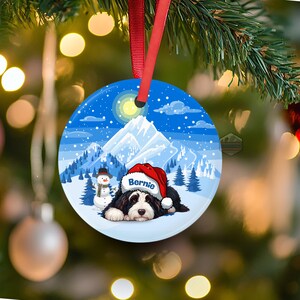 May include: Round Christmas ornament featuring a black and white dog wearing a Santa hat with the name "Bernie". The ornament has a winter scene with a snowman, mountains, and a blue sky.