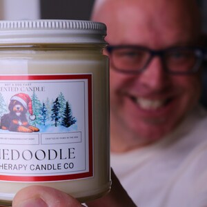 Puede incluir: Vela de aromaterapia en un tarro de cristal con tapa blanca. La etiqueta presenta un perro Bernedoodle con un gorro de Pap&aacute; Noel, con el texto "BERNEDOODLE AROMATHERAPY CANDLE CO." y "SCENTED CANDLE".