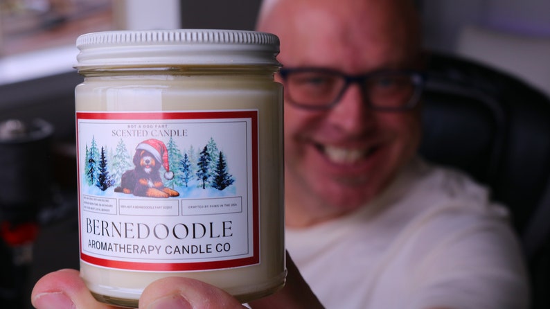 Puede incluir: Vela de aromaterapia en un tarro de cristal con tapa blanca. La etiqueta presenta un perro Bernedoodle con un gorro de Pap&aacute; Noel, con el texto "BERNEDOODLE AROMATHERAPY CANDLE CO." y "SCENTED CANDLE".