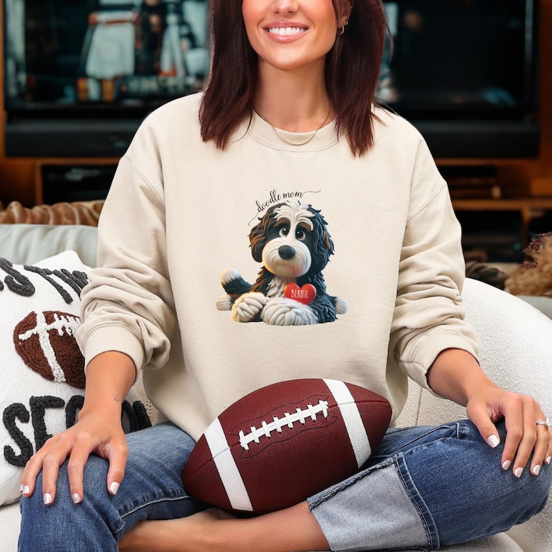 A woman wearing a Bernedoodle sweatshirt with cute design on the front and the back.  The front features a bernedoodle dog in claymation design holding a heart with his name, across the top of his head the words, "Doodle Mom".