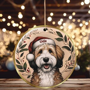 May include: Round Christmas ornament featuring a smiling dog wearing a Santa hat with the name "Otis". The ornament has a gold border and is decorated with a wreath of green leaves and red berries. The background is a bokeh of warm lights.