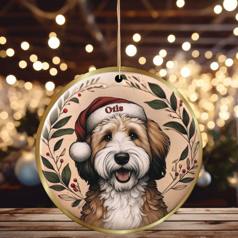 May include: Round Christmas ornament featuring a smiling dog wearing a Santa hat with the name "Otis". The ornament has a gold border and is decorated with a wreath of green leaves and red berries. The background is a bokeh of warm lights.