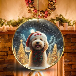 May include: Round Christmas ornament featuring a fluffy dog wearing a Santa hat with the name "Goldie" on it. The dog is wearing a festive green and red scarf. The background shows a snowy winter scene with Christmas trees and a full moon.