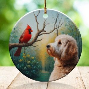 May include: Round ceramic ornament featuring a red cardinal perched on a tree branch, facing a fluffy, light-brown dog. The background depicts a forest scene with a path. A gold string is attached for hanging.