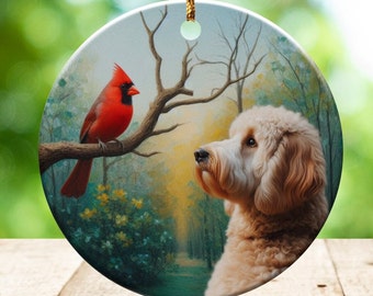 May include: Round ceramic ornament featuring a red cardinal perched on a tree branch, facing a fluffy, light-brown dog. The background depicts a forest scene with a path. A gold string is attached for hanging.