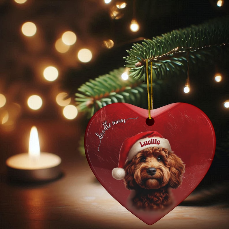 May include: Red heart-shaped Christmas ornament featuring a brown Doodle dog wearing a Santa hat with the name "Lucille". The ornament has the words "doodle mom" and is hanging from a gold string.