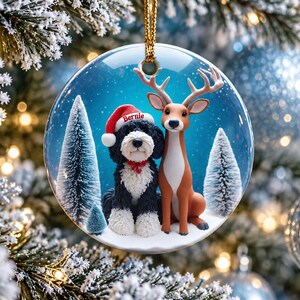 May include: A round ceramic Christmas ornament featuring a black and white dog wearing a red Santa hat with the name Bernie printed on it, sitting next to a brown deer in a snowy forest setting with pine trees under a blue sky.