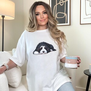 May include: White t-shirt featuring a graphic of a black and white dog. The dog is lying down with its head resting on its paws. The shirt is a relaxed fit. The person is holding a mug with blue, pink, and white stripes.